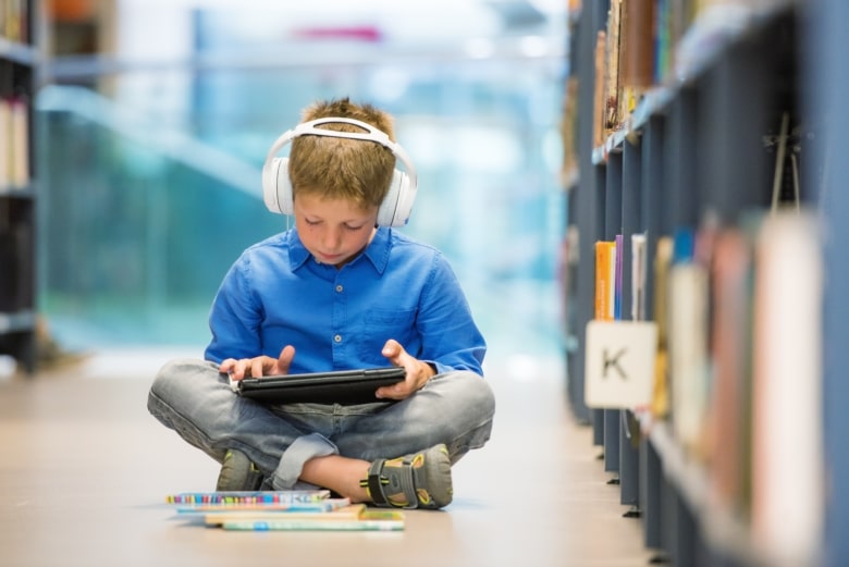 Schoolboy with headphones and digital tablet sitting on the floor in library.